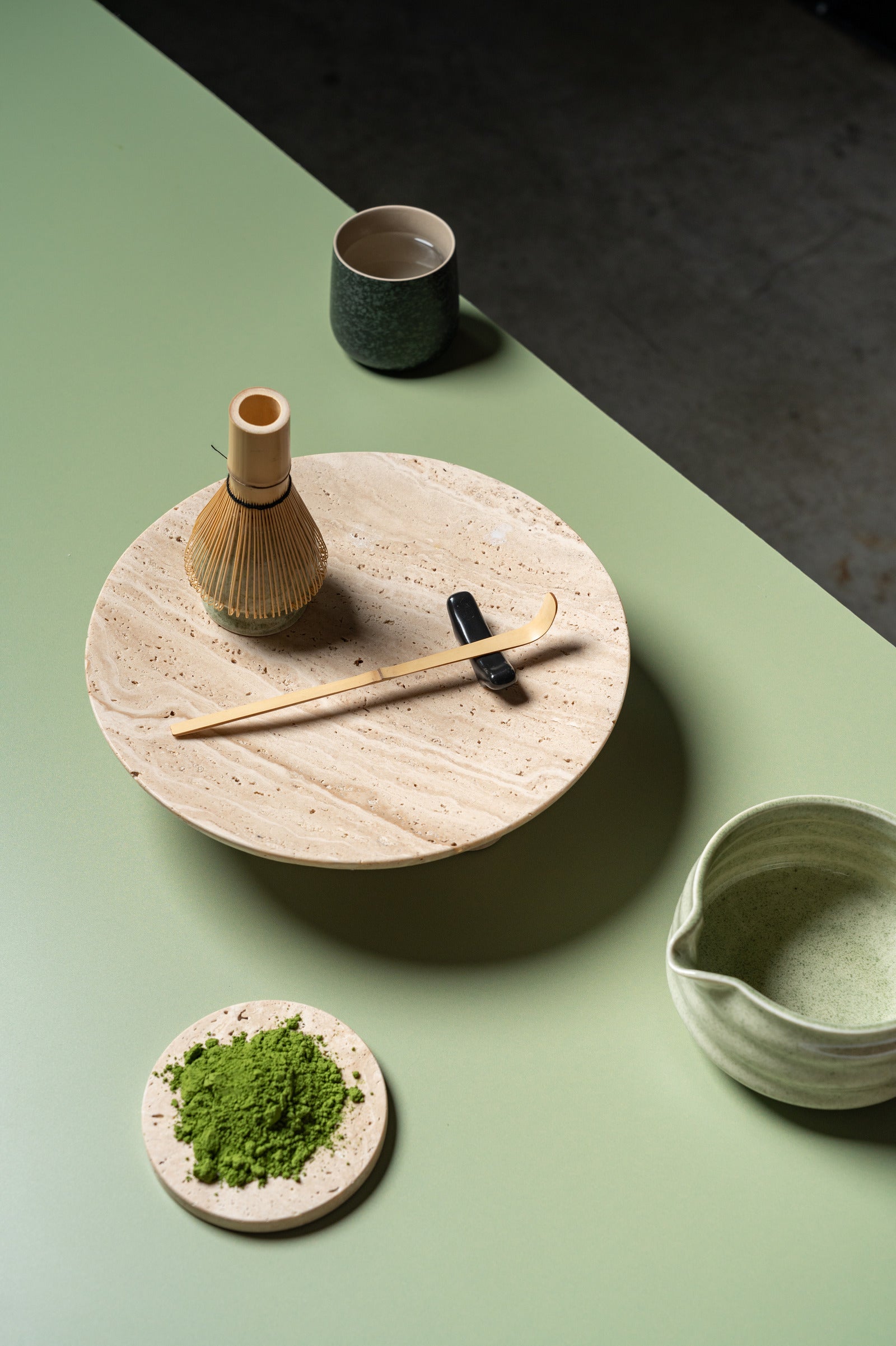 Tea-making setup with a wooden whisk, green tea, and ceramic containers on a green surface.